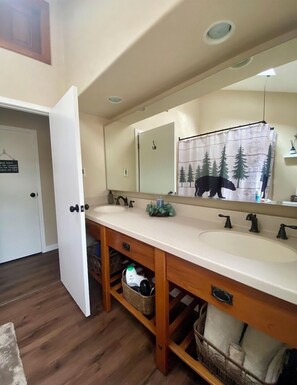 Une salle de bain avec un meuble-lavabo en bois, un grand miroir, un rideau de douche orné d’un motif de forêt et un lavabo blanc.