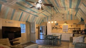 A spacious living room with a television, a dining area with a table and chairs, a kitchen with a refrigerator and microwave, and a ceiling fan.