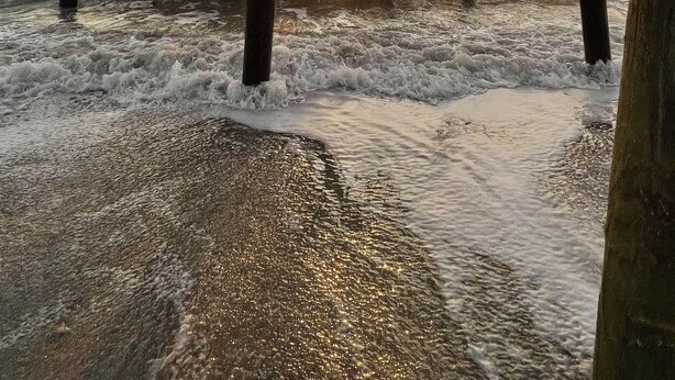 Un muelle con vigas de madera que se extiende sobre el agua, llegando a una playa donde las olas rompen en la orilla.
