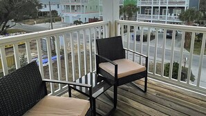 Una terraza de madera con dos sillas de mimbre, baranda blanca y vista a edificios de varios pisos al fondo.