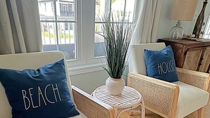 Una habitación con dos sofás blancos, cada uno con una almohada azul, una mesita blanca con una planta y vistas a una casa a través de amplios ventanales.