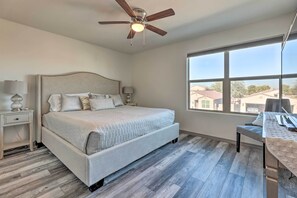 Quarto com cama, escrivaninha, cadeira, ventoinha no teto e uma janela grande.