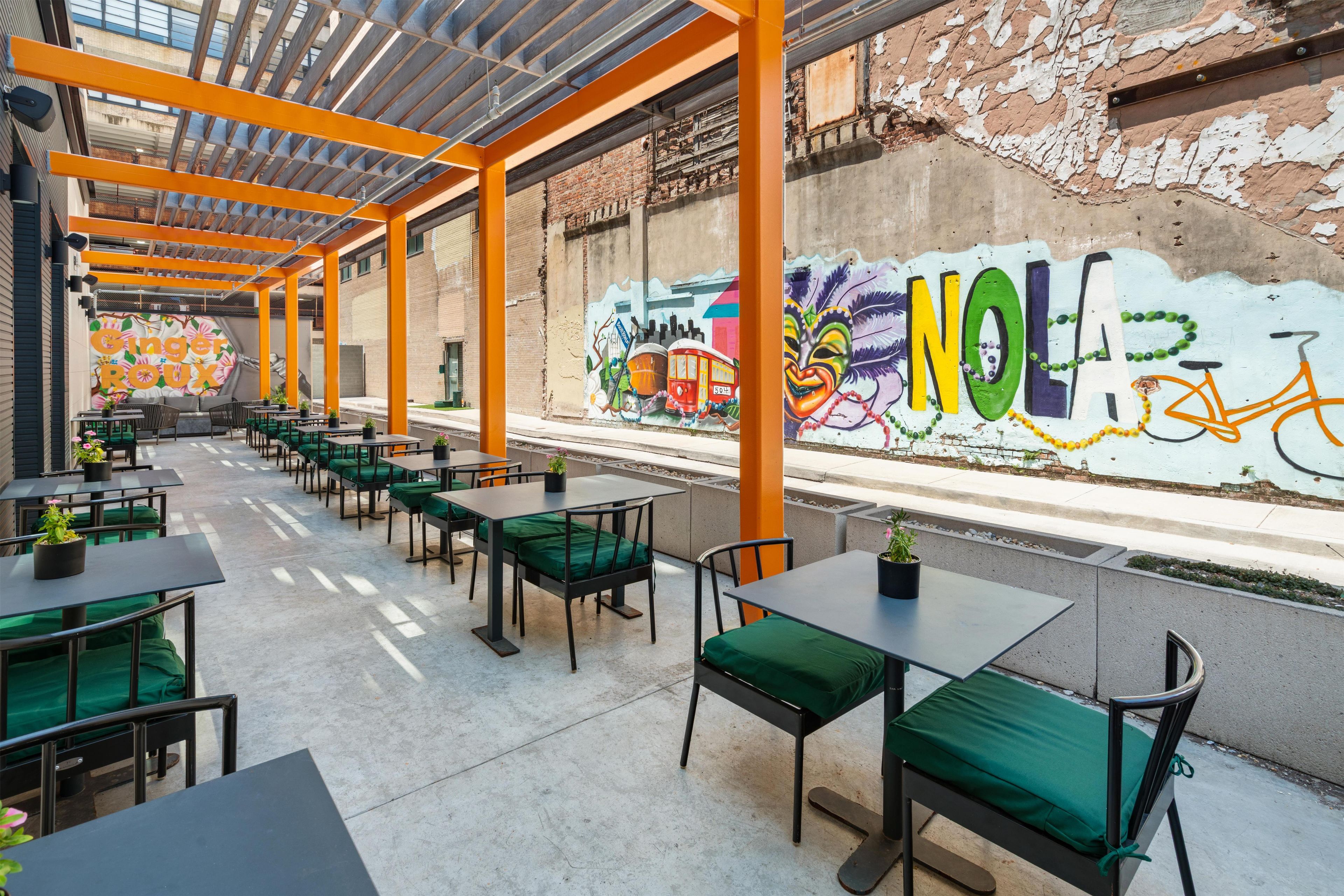 An outdoor café with tables and chairs, a mural on the wall, and a view of the street.
