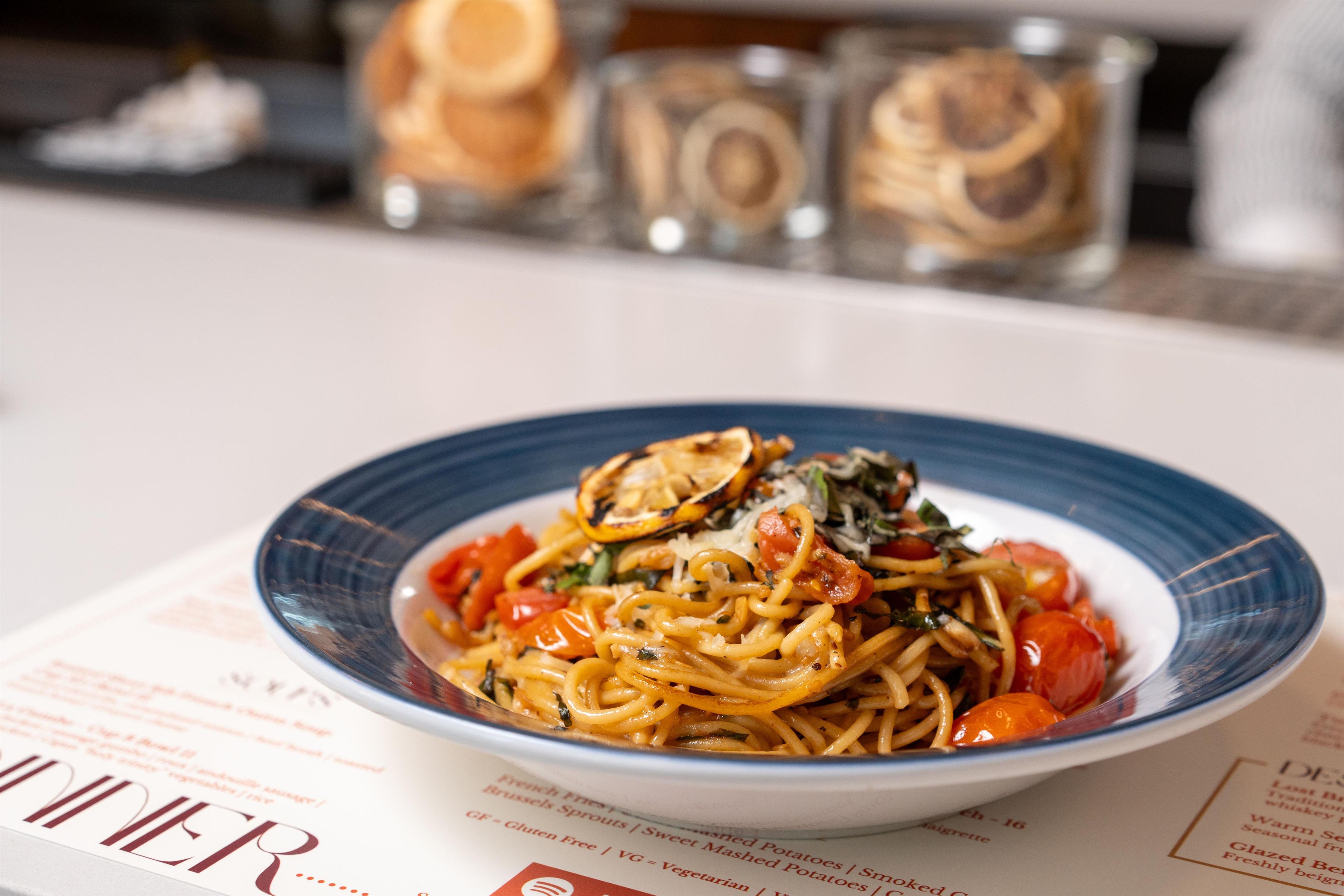 Een bord spaghetti met tomaten en kruiden, geserveerd op een bord met blauwe rand.