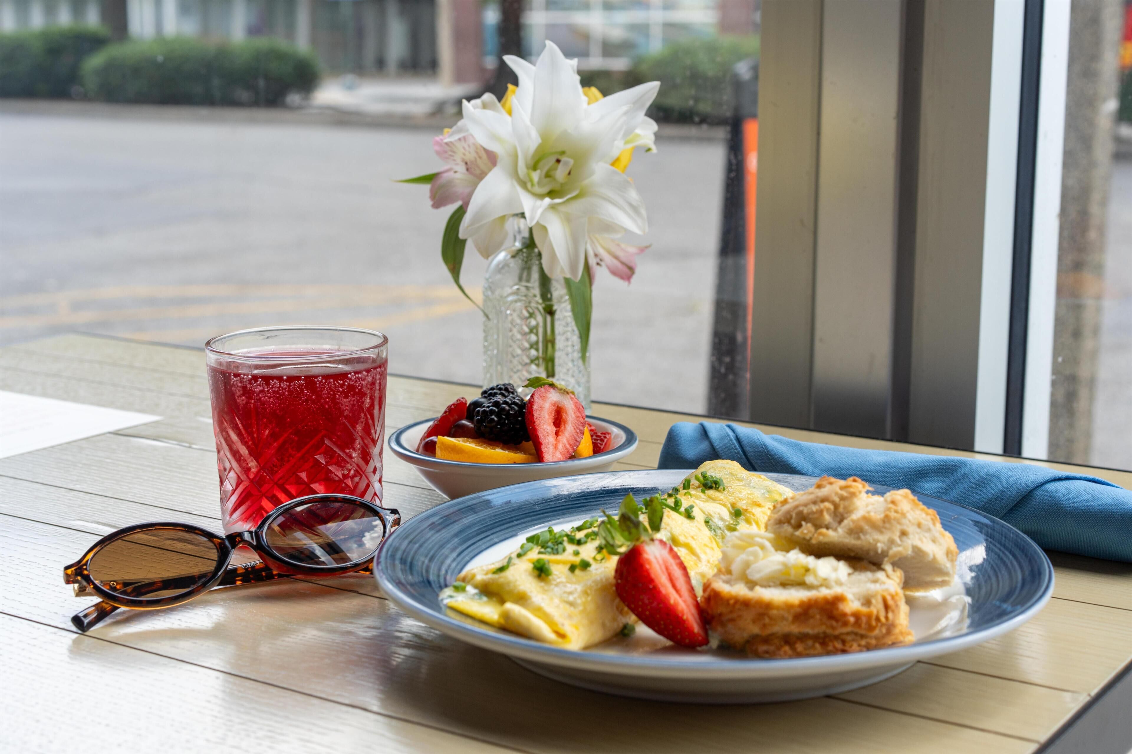 A table with a plate of food, a glass of red beverage, a vase with flowers, and a pair of sunglasses.