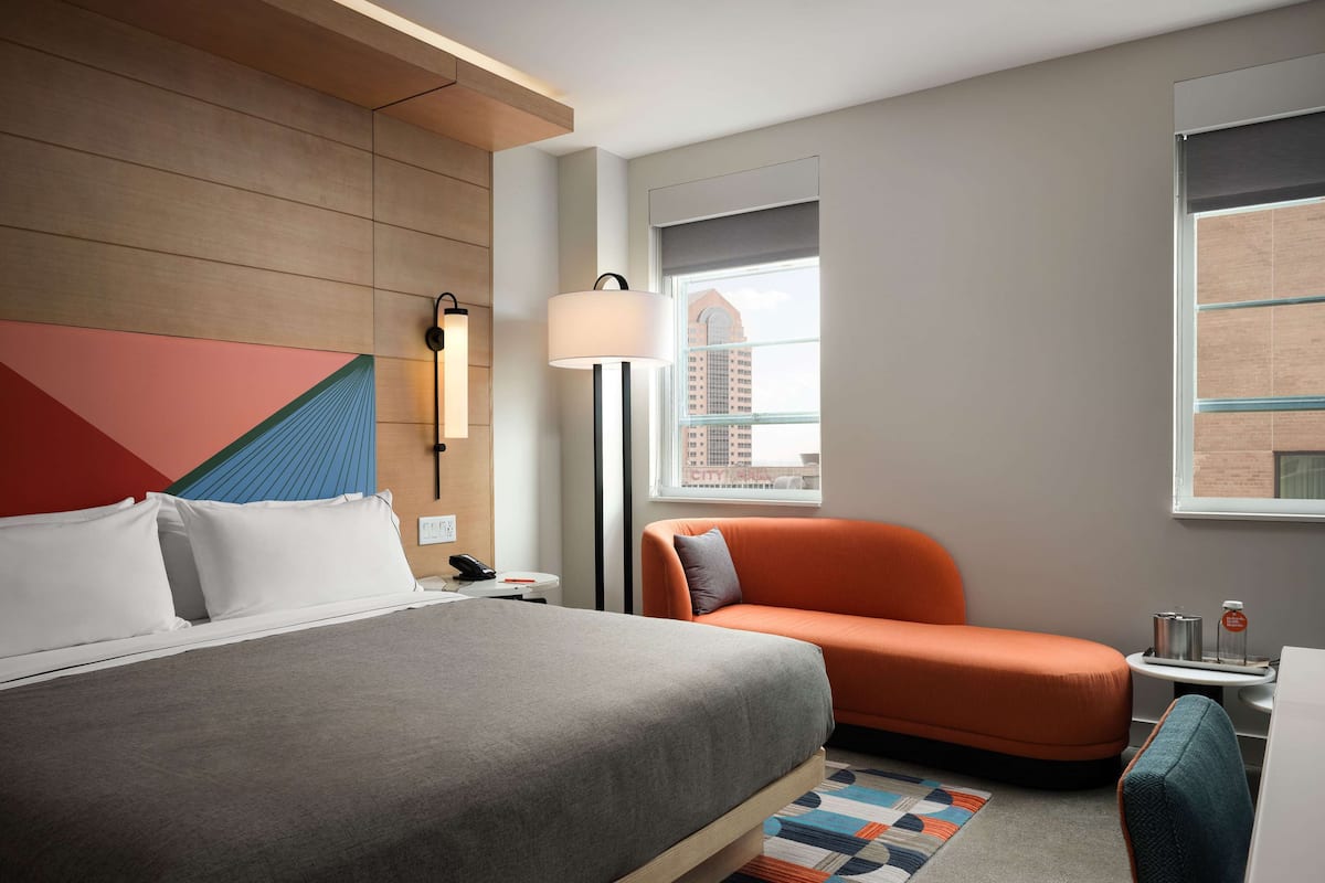 Habitación de hotel con cama, sofá, mesita de noche con lámpara y vistas a la ciudad.
