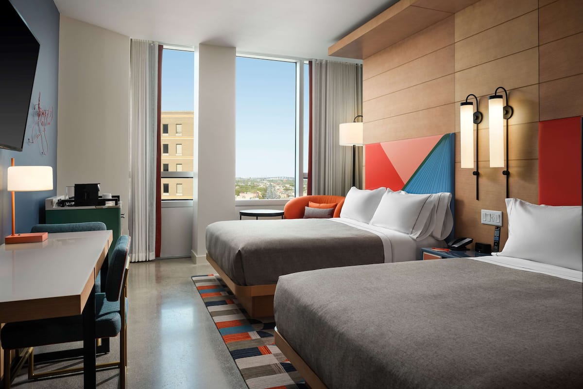 Habitación de hotel con dos camas, un escritorio, una silla, un televisor y vistas a la ciudad.