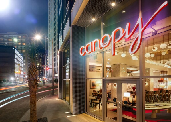 Un edificio moderno con fachada de vidrio y un llamativo letrero con forma de 'canopy'. En el interior, hay varias zonas de estar con mesas y sillas.