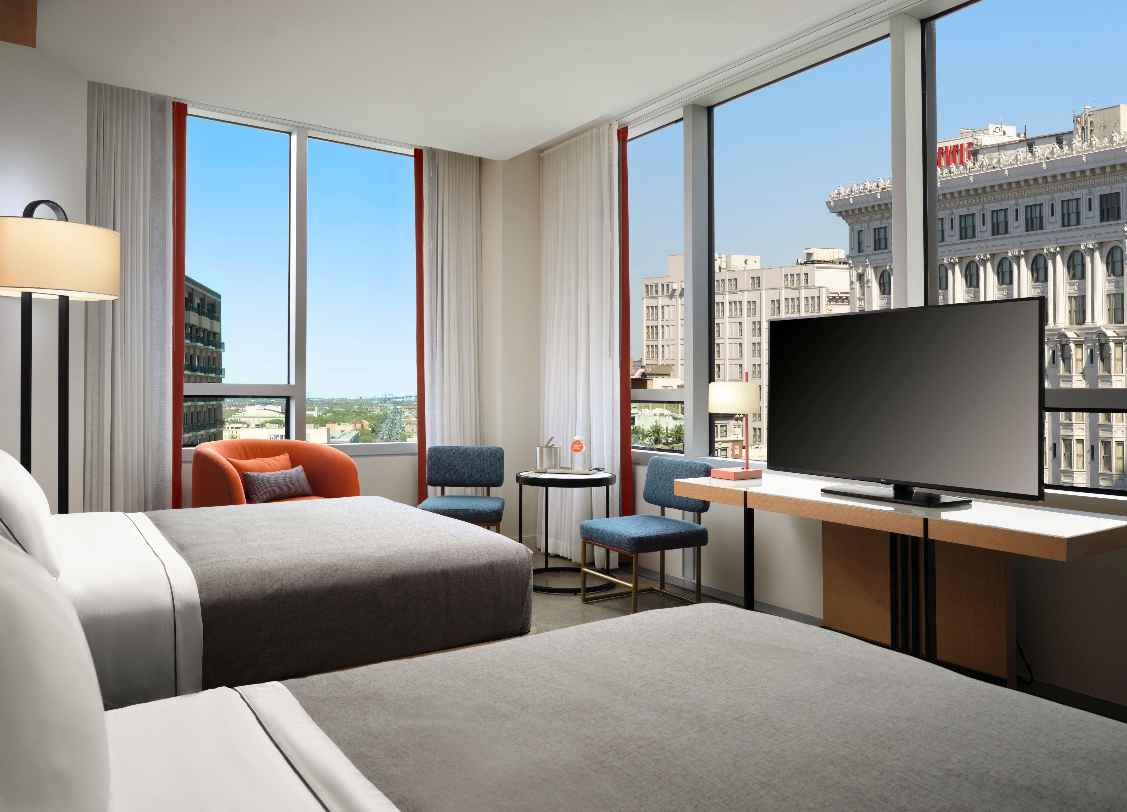 A hotel room with two beds, a desk with a TV, a chair, a small table, and large windows offering a city view.