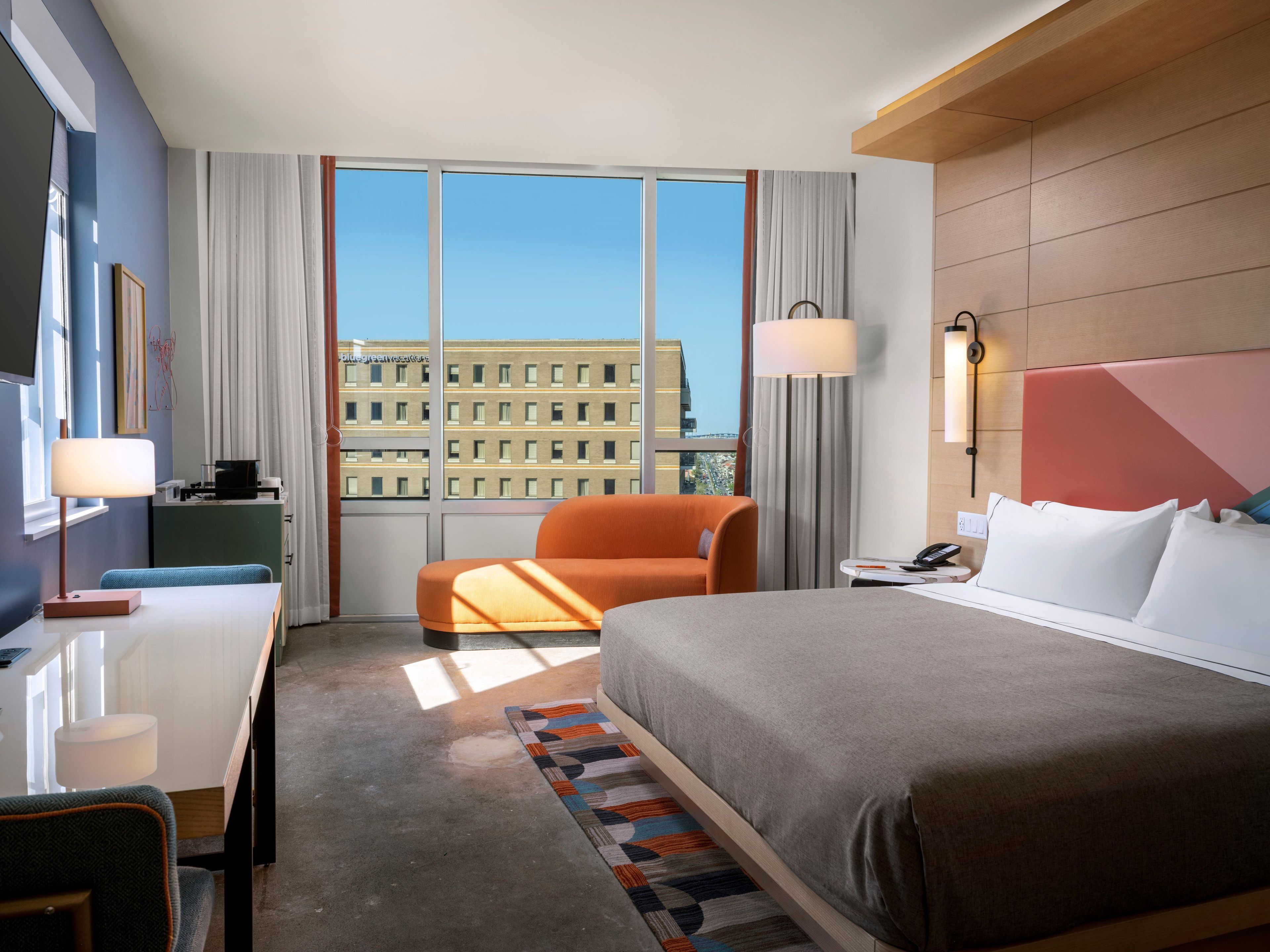Habitación de hotel con cama, escritorio, silla, mesita de noche y vistas a un edificio a través de amplias ventanas.