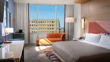 Habitación de hotel con cama, escritorio, silla, mesita de noche y vistas a un edificio a través de amplias ventanas.