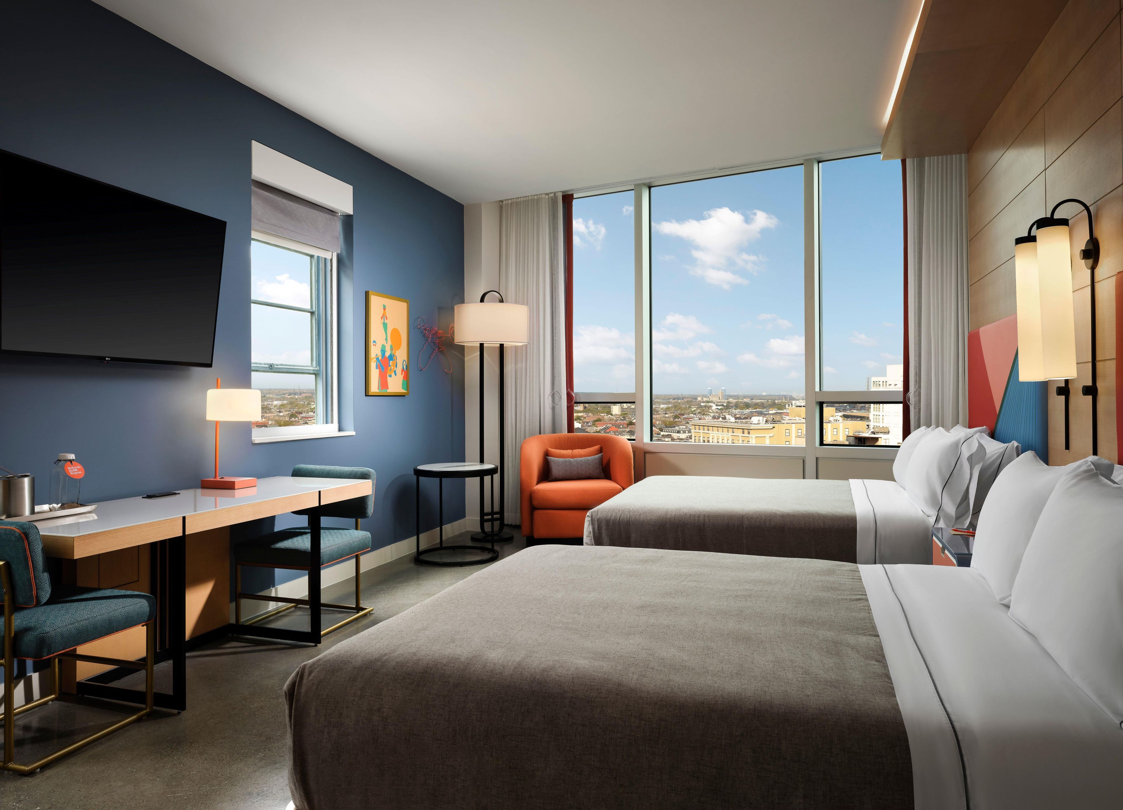 A hotel room with a bed, a desk, a chair, a sofa, a TV, and a large window with a city view.