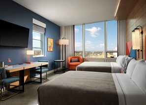 Ein Hotelzimmer mit einem Bett, einem Schreibtisch, einem Stuhl, einer Couch, einem Fernseher und einem großen Fenster mit Blick auf die Stadt.
