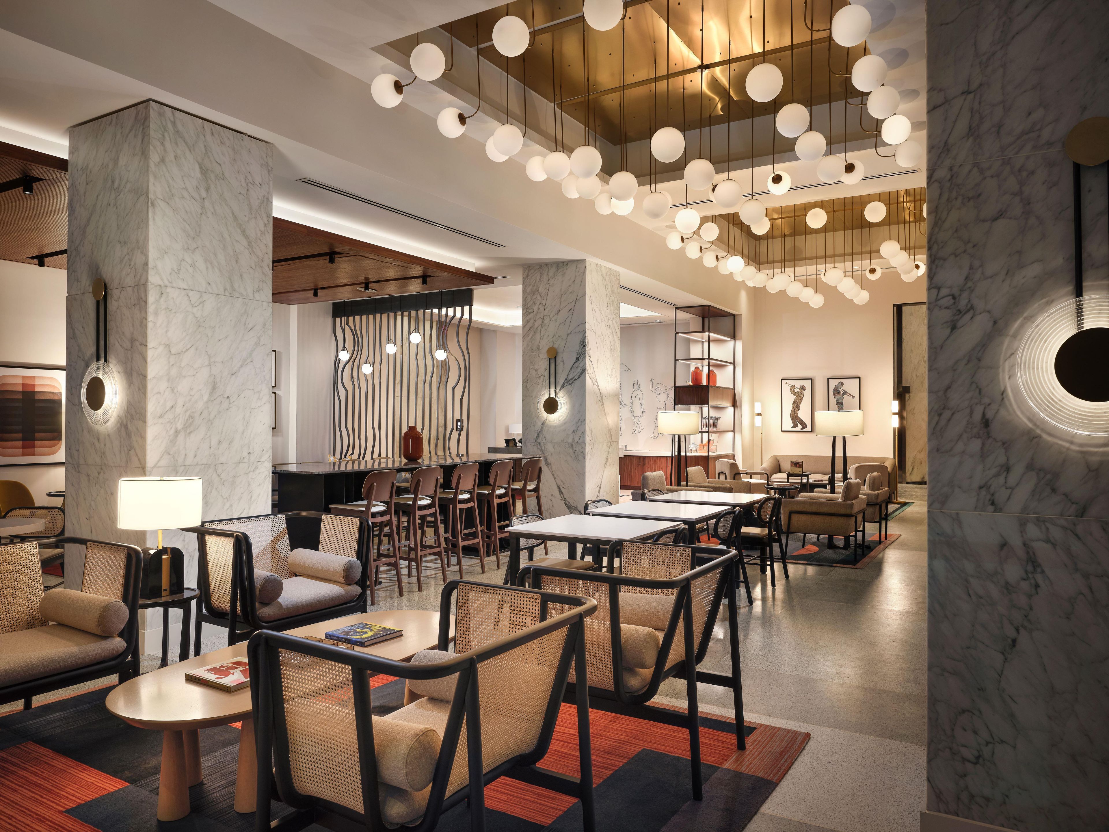 A modern hotel lobby with marble columns, a central bar area, and a seating arrangement with tables and chairs.