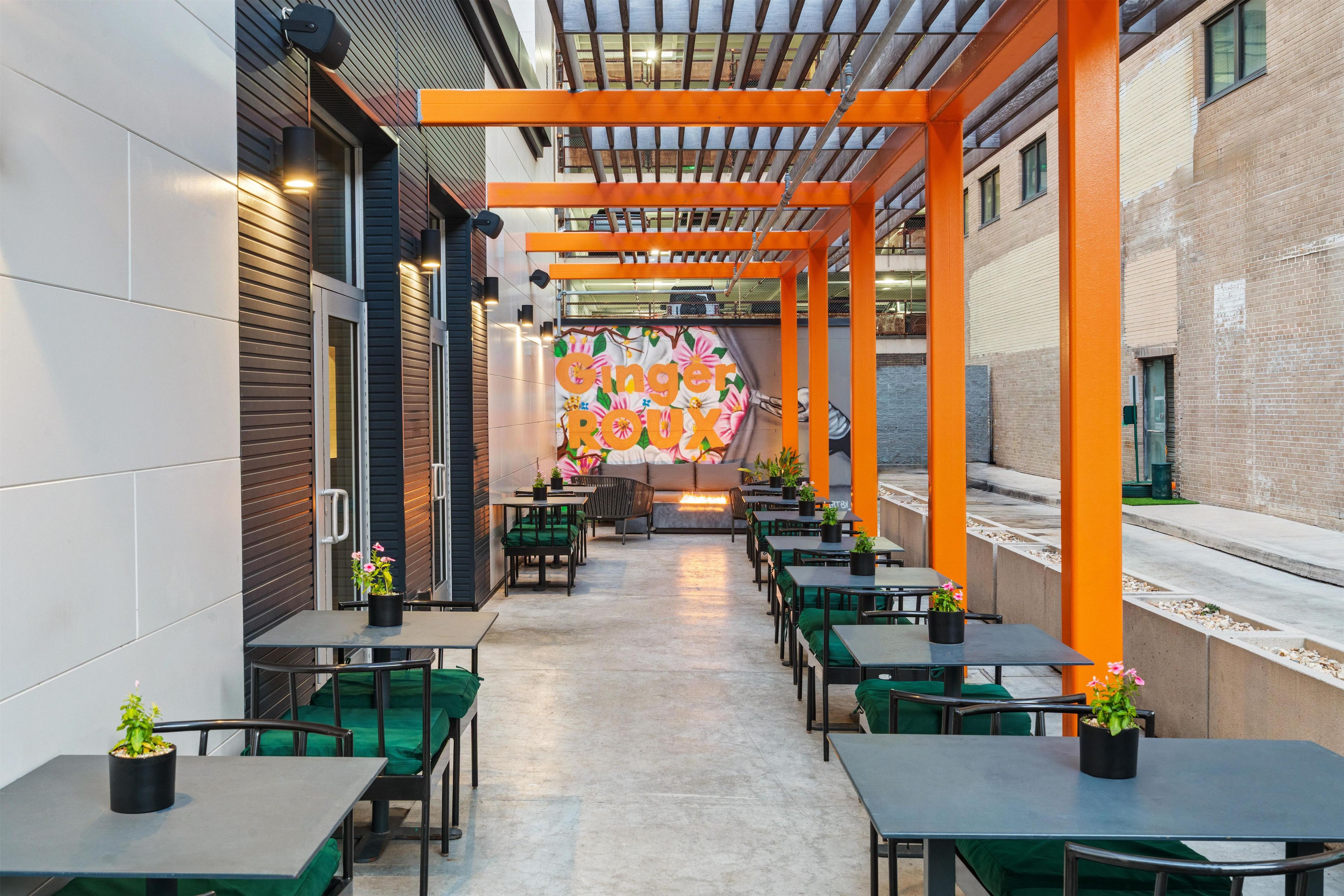 An outdoor dining area with tables and chairs, a colorful mural, and a brick building in the background.