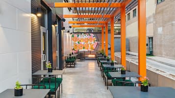 An outdoor dining area with tables and chairs, a colorful mural, and a brick building in the background.