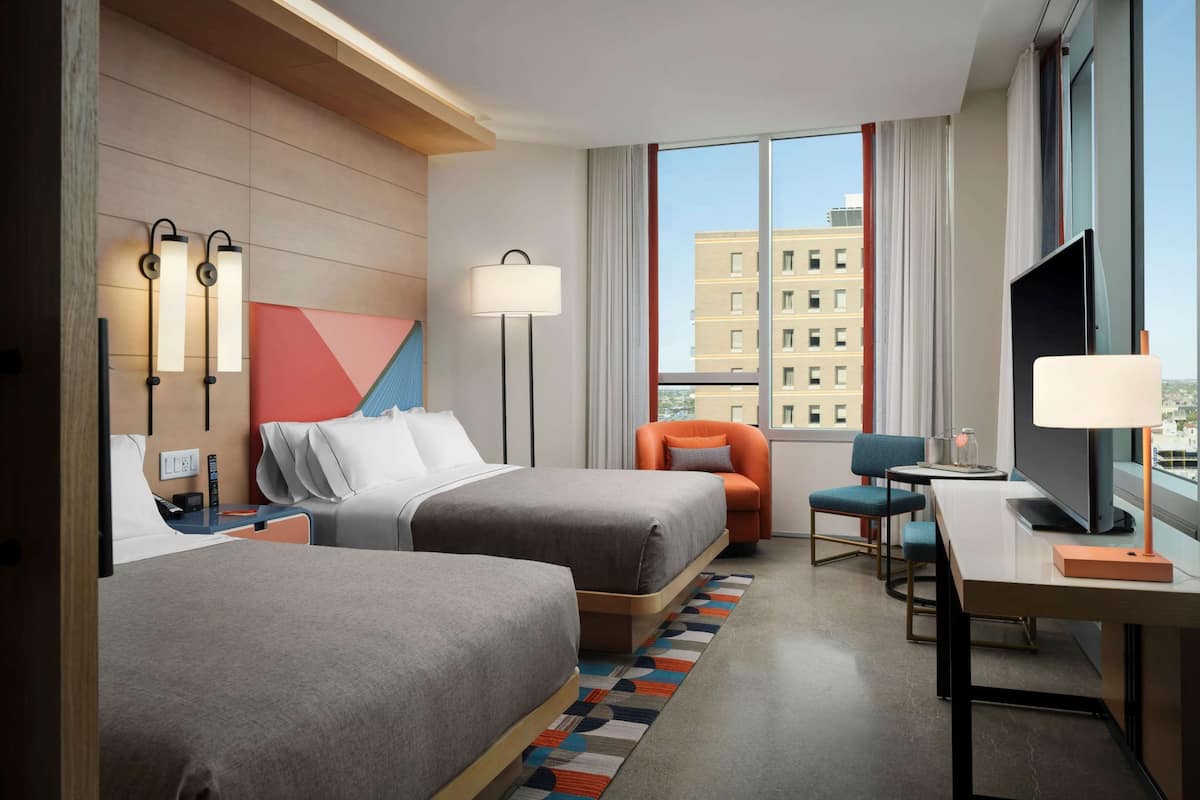 Habitación de hotel con dos camas, un escritorio, una silla, un televisor y un ventanal con vista a la ciudad.