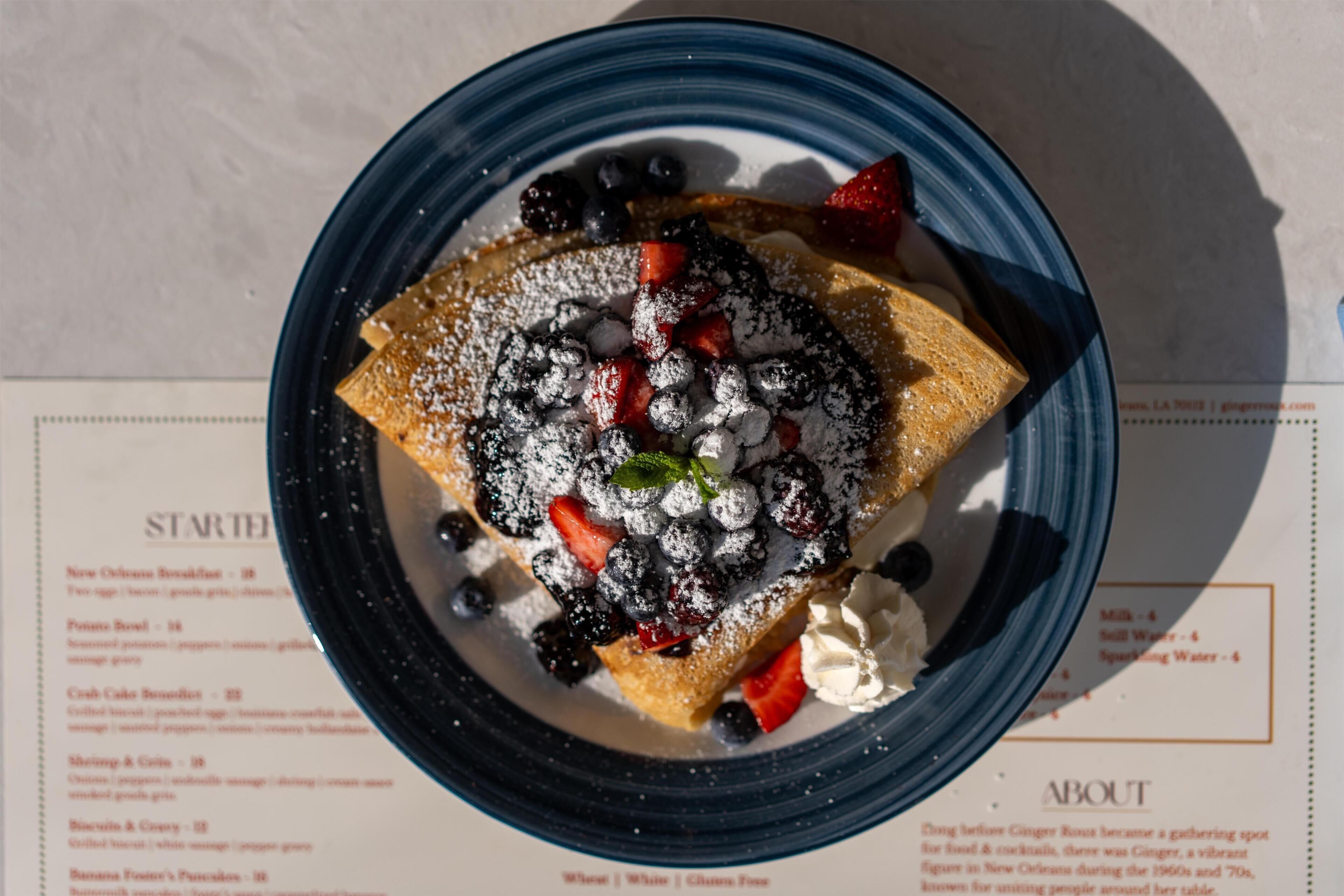 A dessert plate with a crepe, berries, and powdered sugar on a table with a menu.