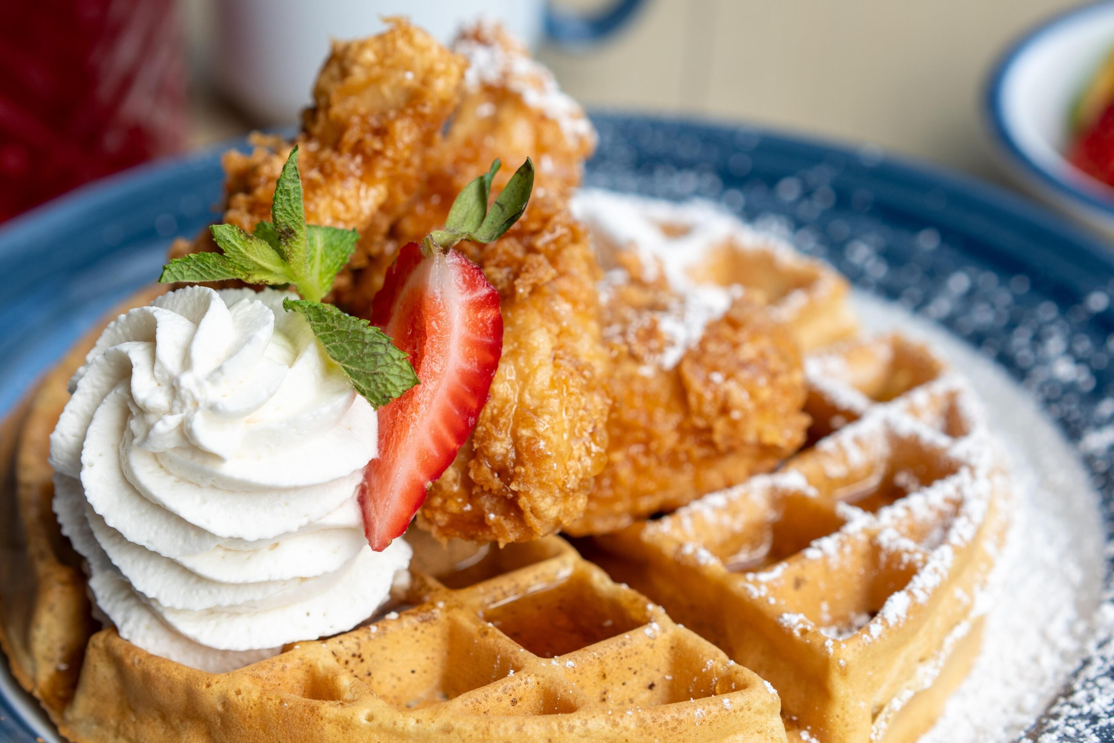 A plate of waffles with whipped cream and a strawberry, accompanied by a piece of fried chicken.