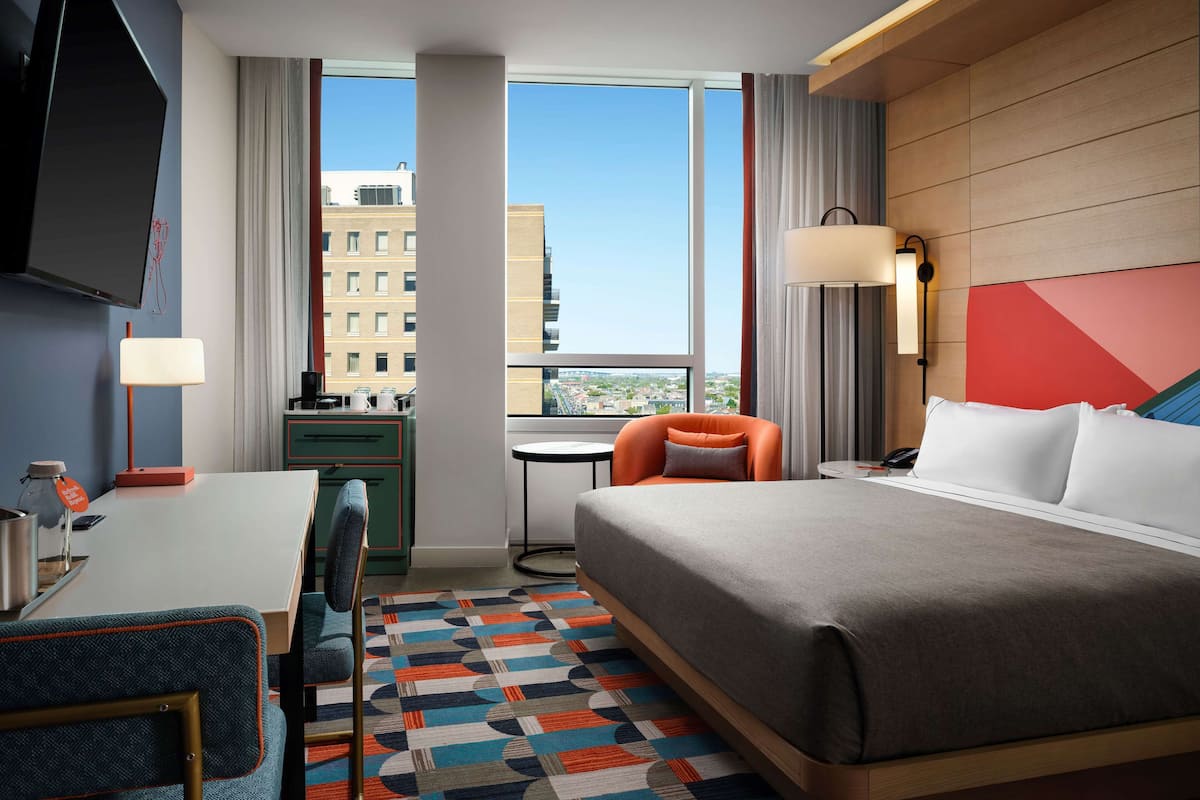Habitación de hotel con cama, escritorio, silla, televisión y vistas a edificios.