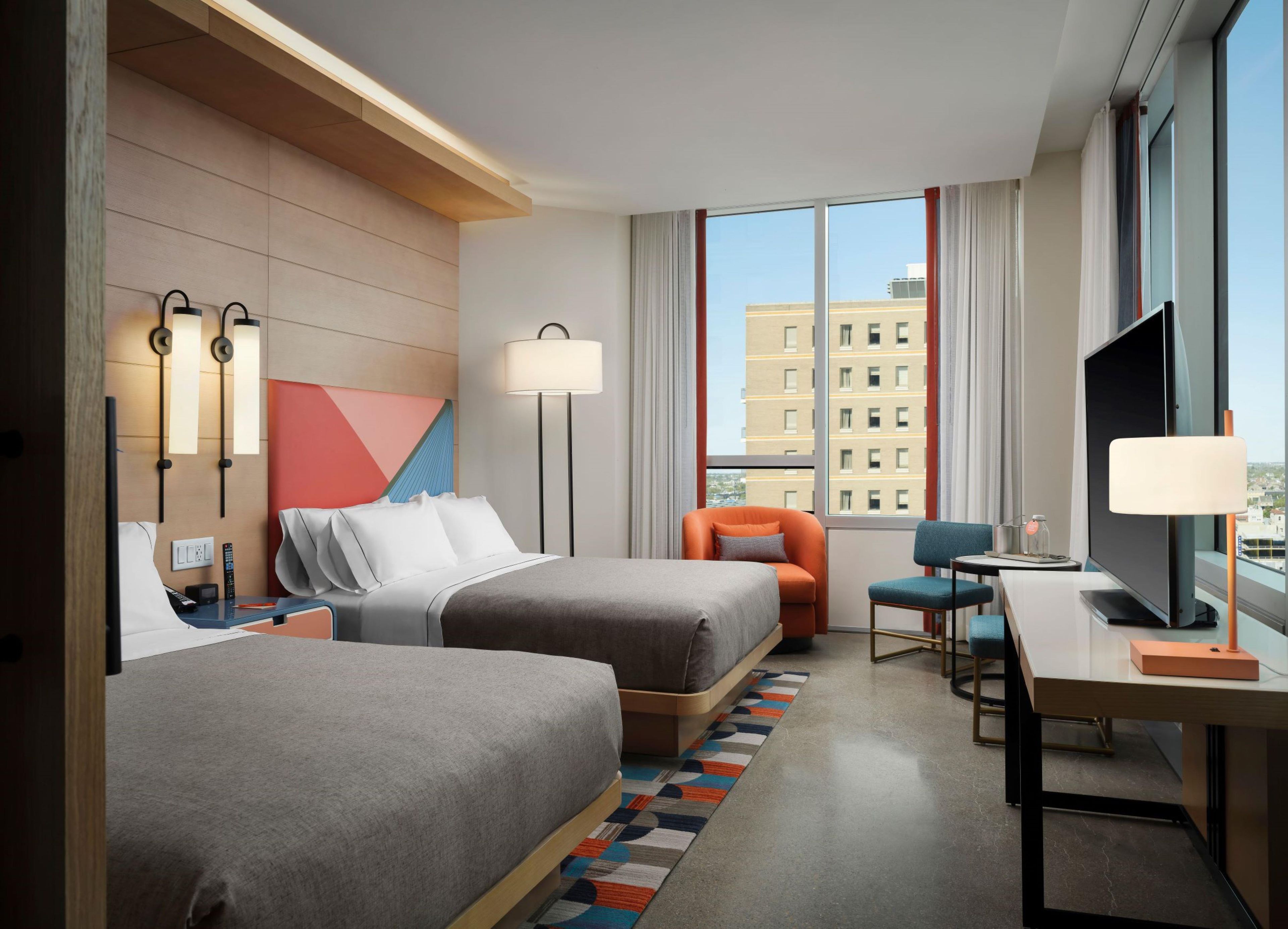 A hotel room with two beds, a desk, a chair, a TV, and a large window with a city view.