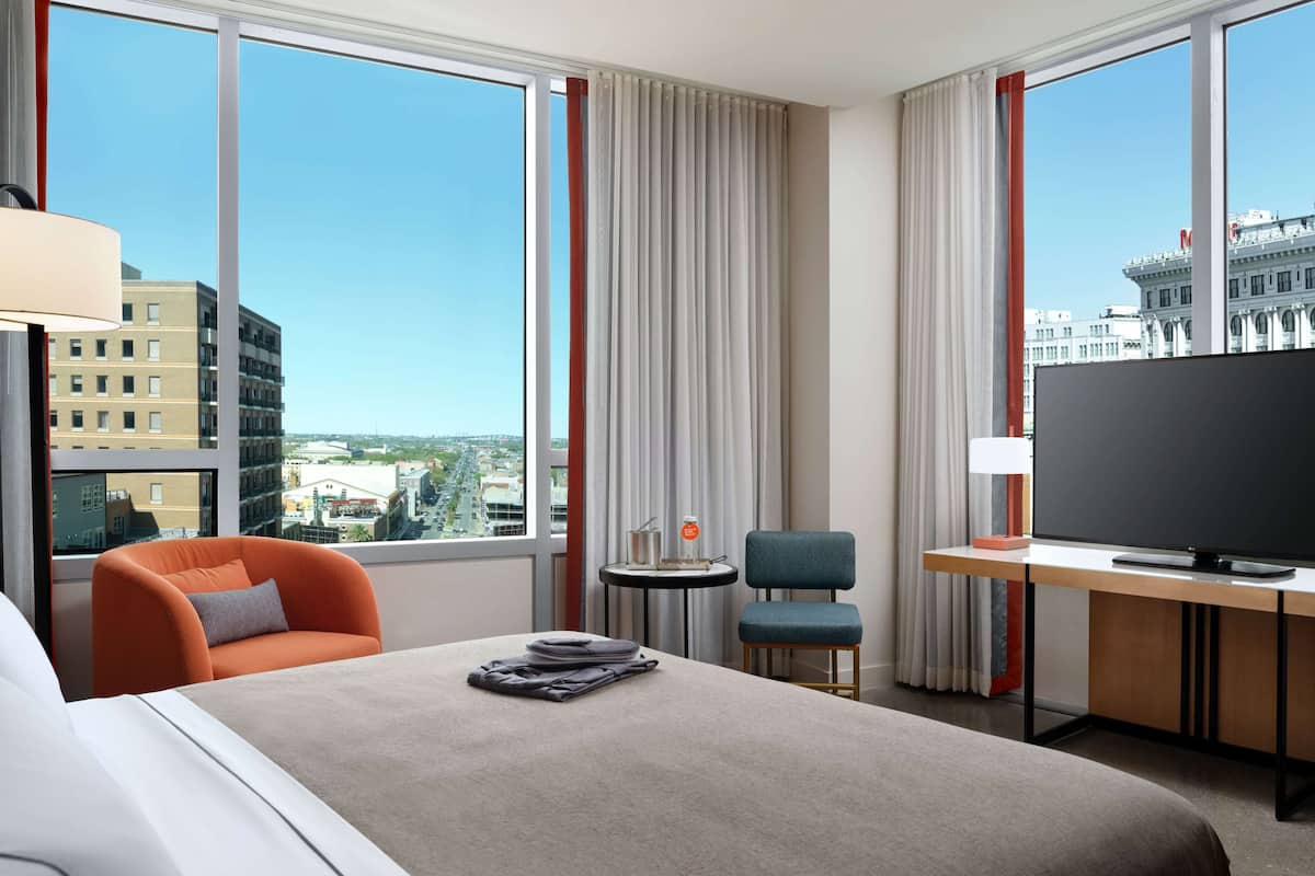 Habitación de hotel con cama, escritorio, silla, televisor y ventanas amplias que ofrecen vistas de la ciudad.