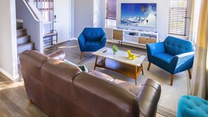 Ein Wohnzimmer mit einem braunen Ledersofa, zwei blauen Sesseln, einem Holztisch und einem Fernseher auf einem weißen Schrank.
