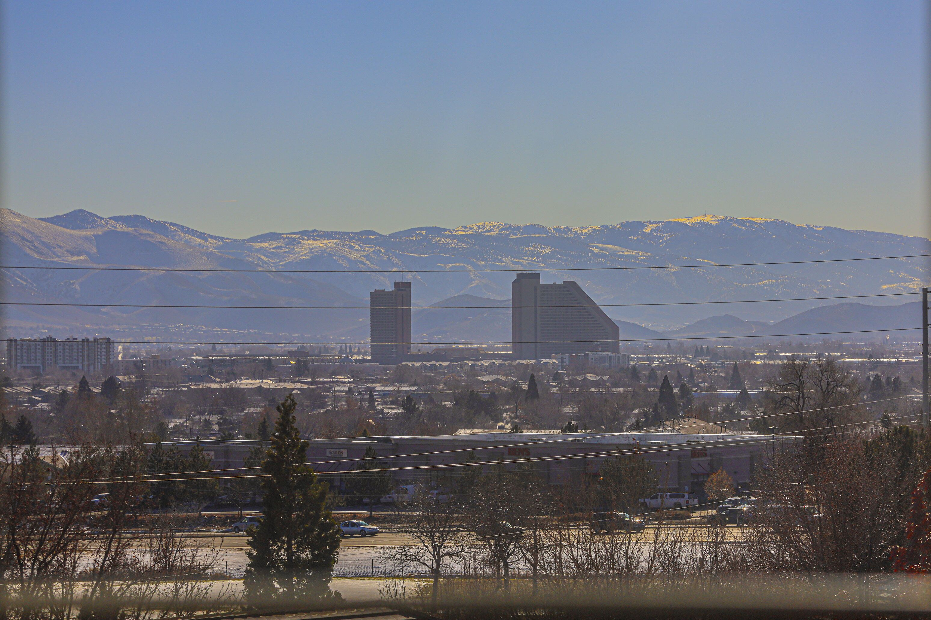A cityscape with snow-capped mountains in the background, a parking lot, and several buildings including a tall structure.