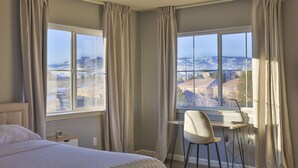 Ein Hotelzimmer mit Bett, Schreibtisch und Stuhl. Große Fenster mit Vorhängen bieten einen Blick auf die Stadtlandschaft.