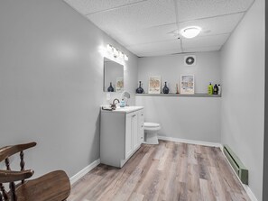 Ein modernes Badezimmer mit Holzboden, einem weißen Waschbecken und einer Toilette. An den Wänden hängen Bilder in Rahmen und über dem Waschbecken befindet sich ein Spiegel.