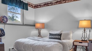 A neatly made bed with a quilted white comforter, two bedside lamps, a wall-mounted fan, and a window with a view of trees.
