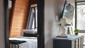 A bedroom with a sloped ceiling, a bed with white bedding, a bedside table with a bust sculpture, and a window with a view of trees. - Vintage Waterfront A Frame- Hideaway On The Bay (Lusby)