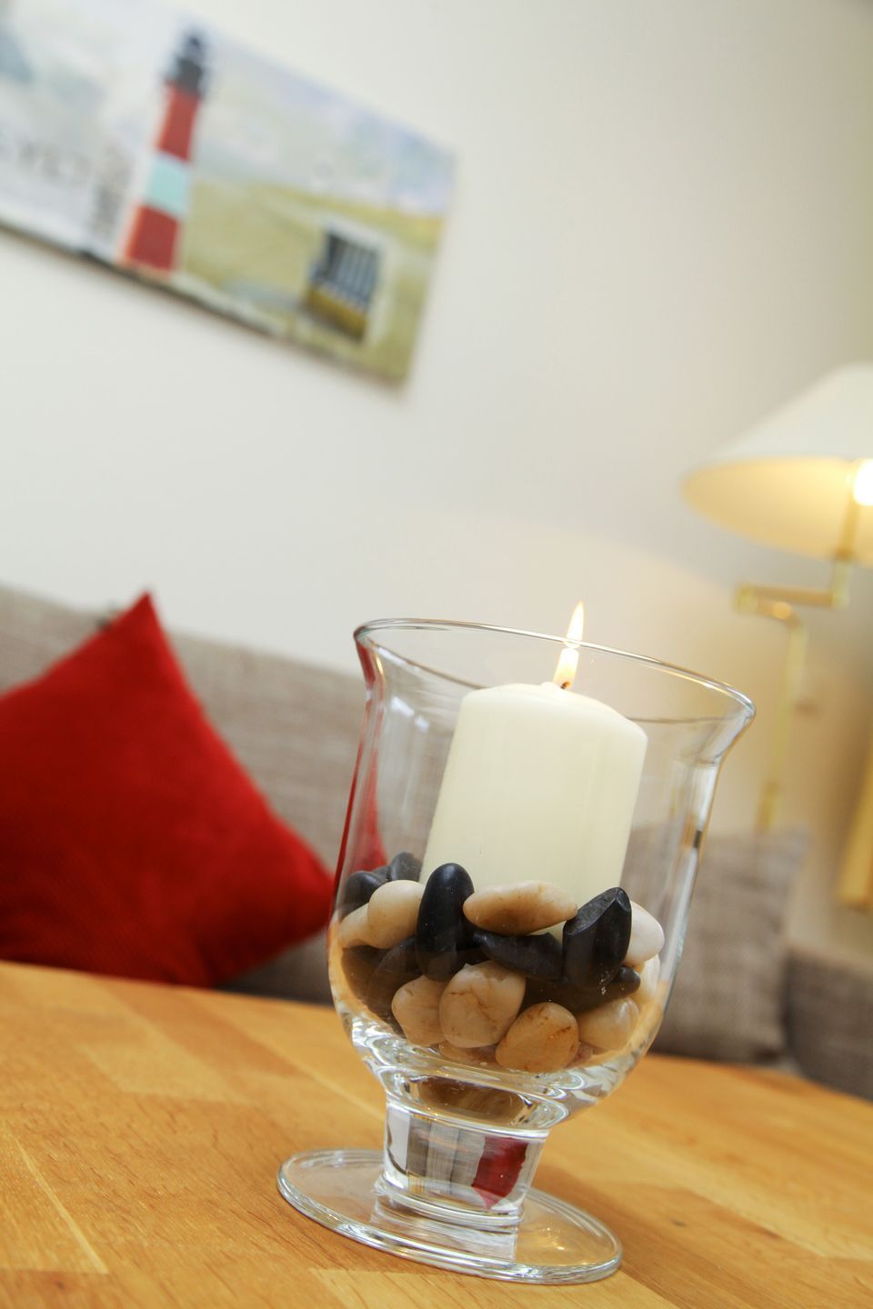 A candle in a glass holder on a wooden table with a red pillow in the background.