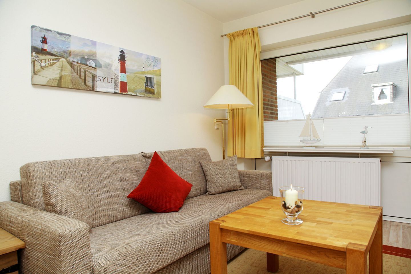 A cozy living room with a sofa, a wooden coffee table, a painting of a lighthouse, a window with curtains, and a radiator.