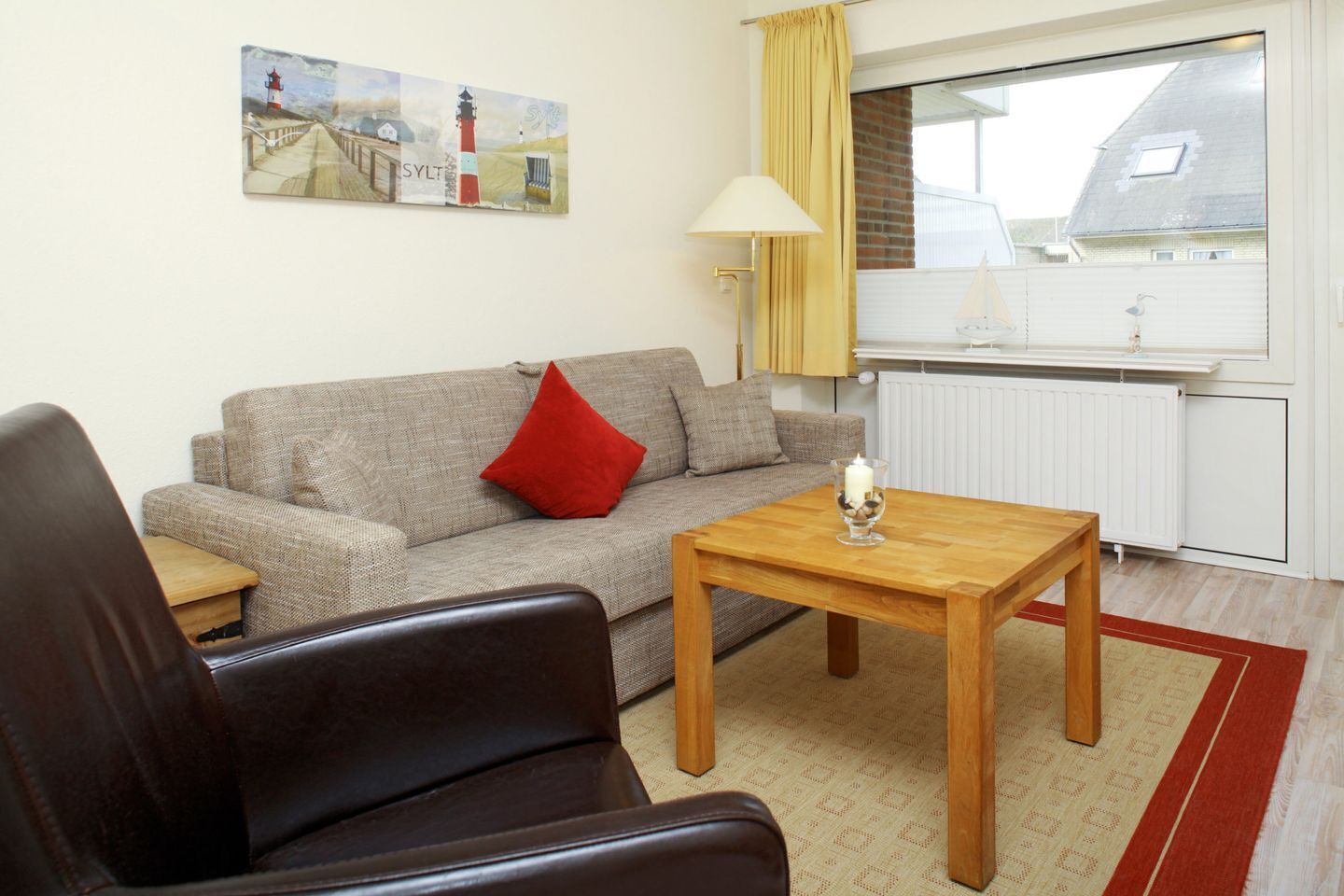 Un salon comprenant un canapé, une table basse en bois, un coussin rouge, une lampe et un tableau représentant des phares.