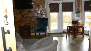 Una sala de estar con pared de piedra, un sillón azul, un sofá gris, una chimenea y una zona de cocina con taburetes de bar.