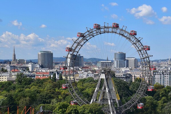A large Ferris wheel with gondolas, surrounded by a cityscape with various buildings and a prominent tower in the background.