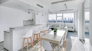Een moderne keuken met een centraal eiland, witte barkrukken, een eettafel met stoelen en grote ramen met uitzicht over de stad.
