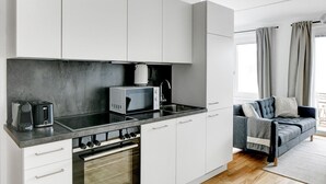 Une cuisine moderne avec des meubles blancs, un four à micro-ondes, une bouilloire et un grille-pain. Elle est équipée d'un four encastré et d'un évier. La pièce comprend un canapé gris et une fenêtre avec des rideaux.