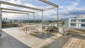 Eine Dachterrasse mit Holzboden, weißer Gartenmöbel und einer Glasüberdachung mit Blick auf die Stadt.