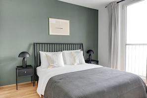 Un dormitorio con una cama, dos mesitas de noche, un tocador, un cuadro enmarcado en la pared y una ventana con cortinas.