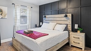 Une chambre à coucher moderne avec un grand lit, une table de chevet, une lampe fixée au mur et une fenêtre avec des rideaux.