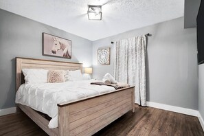 A bedroom with a wooden bed, white bedding, a nightstand with a lamp, a framed picture on the wall, and a window with curtains. - ☆Stunning Derby Home☆ 5 KING BEDS, HOT TUB,FIREPIT (Jeffersonville)