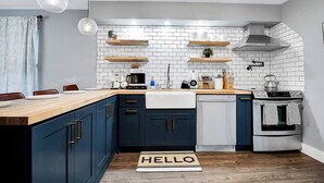 Une cuisine moderne avec des meubles de couleur bleu foncé, des carreaux de céramique blancs et des plans de travail en bois. On y trouve des luminaires suspendus, une machine à café et une bouilloire sur la cuisinière.