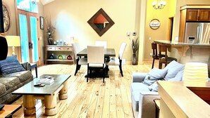 A spacious living room with a wooden floor, a dining area with a table and chairs, a kitchen with wooden cabinets, and a large window with curtains. - Riverbend Retreat with Hot Tub (Traverse City)