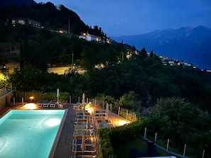 Zona de piscina con sillones de descanso y sombrillas, rodeada de vegetación y con vistas a un paisaje montañoso.
