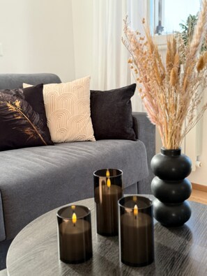 Un salon moderne avec un canapé gris, des coussins à motifs et un vase contenant des plantes séchées. Trois bougies sont posées sur une table ronde.