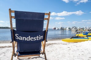 A beach chair with 'Sandestin' written on it, positioned on a sandy beach with boats in the background.