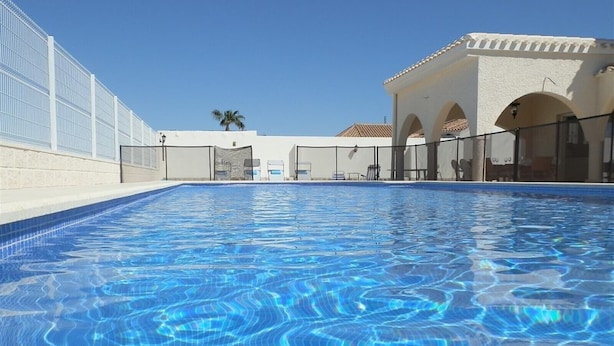 A swimming pool with clear blue water, surrounded by a white building with arches and a palm tree in the background.