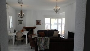 A spacious living room with a dark brown sofa, a white dining table with chairs, and a chandelier.